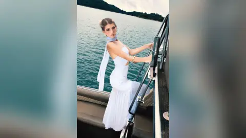 Elegant woman in a white halter gown posing on a luxury boat overlooking a serene blue ocean landscape.