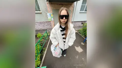 MagalyBoffa wears black sunglasses and a white puffer jacket while standing outside near some green plants and a building.