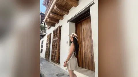 Smiling woman in a beige summer dress and straw hat standing by a rustic wooden door in a Mediterranean village.