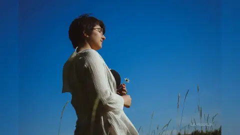 Thoughtful woman in a white linen shirt standing in a sunny meadow under a clear blue sky.