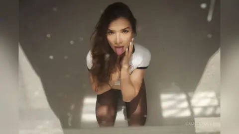 Playful young woman in a white top and sheer black tights posing in a bright, minimalist indoor setting.