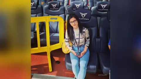 Smiling woman in a patterned long-sleeve shirt and blue jeans sitting in blue stadium seats.