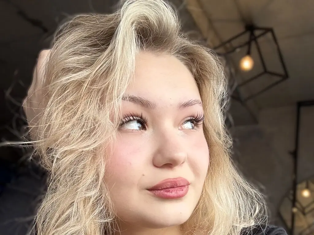 Close-up portrait of a blonde woman with long wavy hair and natural makeup looking upward in soft indoor lighting.
