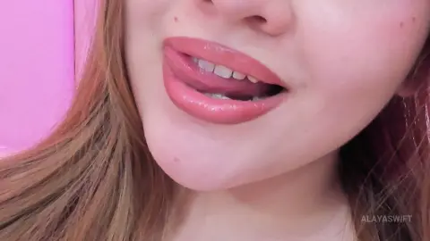 Close-up of a young woman with pink lipstick smiling playfully in a soft pink lit room.
