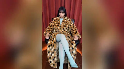 Stylish woman in a leopard print faux fur coat and light blue leggings sitting against a red curtain backdrop.
