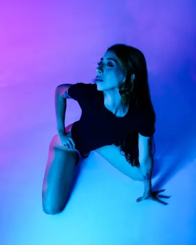AilynHoney — AilynHoney poses on the floor in a black t-shirt under blue and purple neon lights.