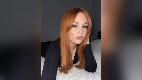 Redheaded woman in a black long-sleeve top posing with a soft, moody expression in a cozy bedroom setting.