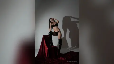Confident woman in black lingerie and tall boots posing on a red velvet draped pedestal in moody studio lighting.