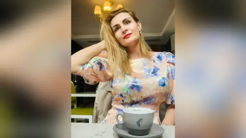 Blonde woman in a floral peach blouse smiling at a cafe table with a cup of coffee.