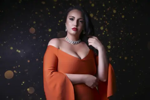 Elegant woman in an orange off-the-shoulder gown and diamond necklace posing against a dark bokeh background.