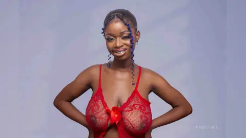Smiling young woman in a red lace bodysuit posing against a soft purple studio background.