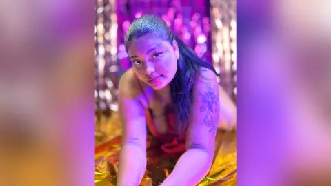 Young woman in red outfit posing against shimmering gold tinsel backdrop with moody purple lighting.