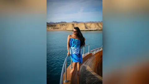 MissyJolie stands on a wooden boat deck wearing a blue patterned tube top and matching skirt, looking out over the blue water.