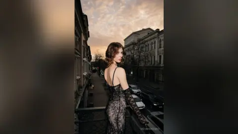 Elegant woman in a black lace evening gown posing on a city balcony during a moody sunset.