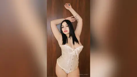 Young woman with long dark hair wearing a cream lace corset bodysuit posing against a wooden wall.