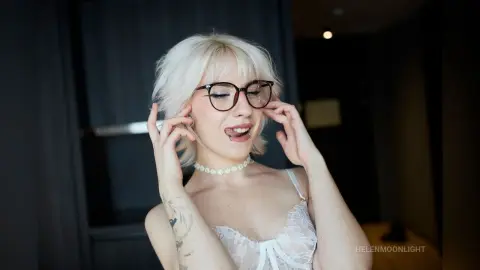 Blonde woman with glasses in white lace lingerie posing playfully in a dark indoor setting.