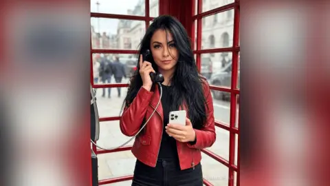 Stylish woman in a red leather jacket talking on a vintage phone inside a classic red London telephone booth.