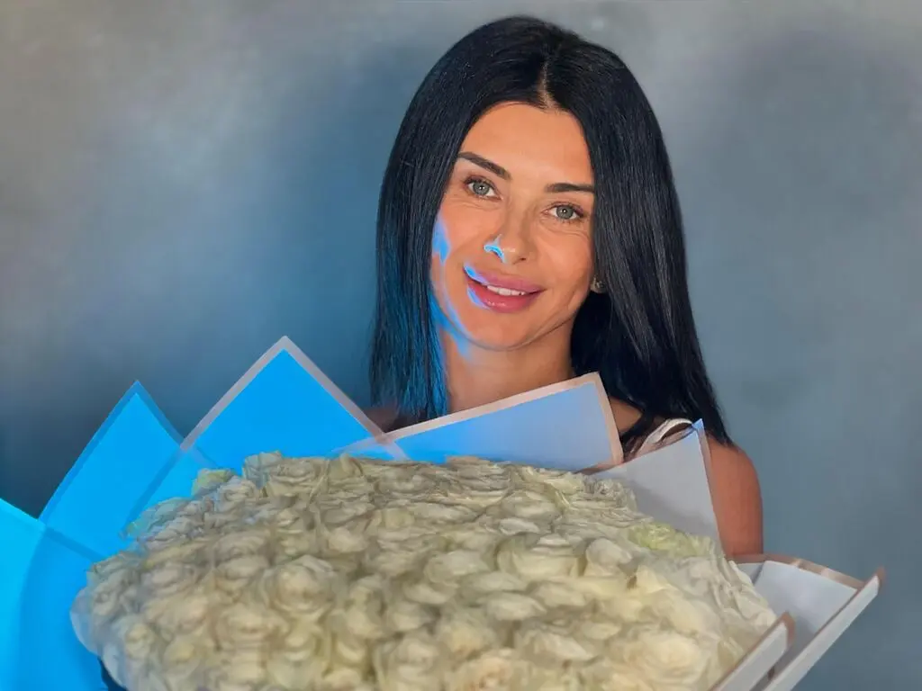 Smiling brunette woman holding a large bouquet of white roses against a soft blue background.