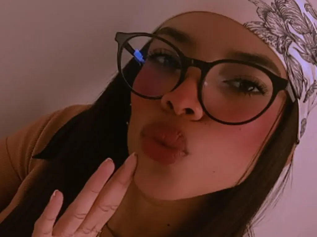 Young woman with glasses making a playful kissy face in warm indoor lighting.