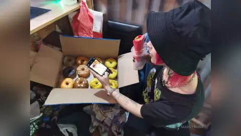 Young woman in a black bucket hat holding a smartphone while looking at a box of assorted donuts indoors.