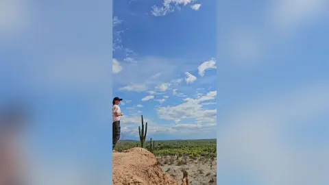 RossiNoleen stands on a dirt ledge overlooking a desert landscape under a blue sky with light clouds.