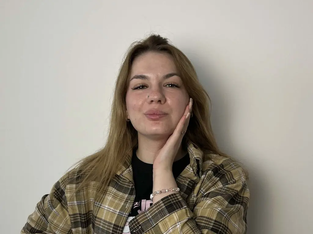 Smiling young woman with long blonde hair wearing a brown plaid flannel shirt against a plain white wall.