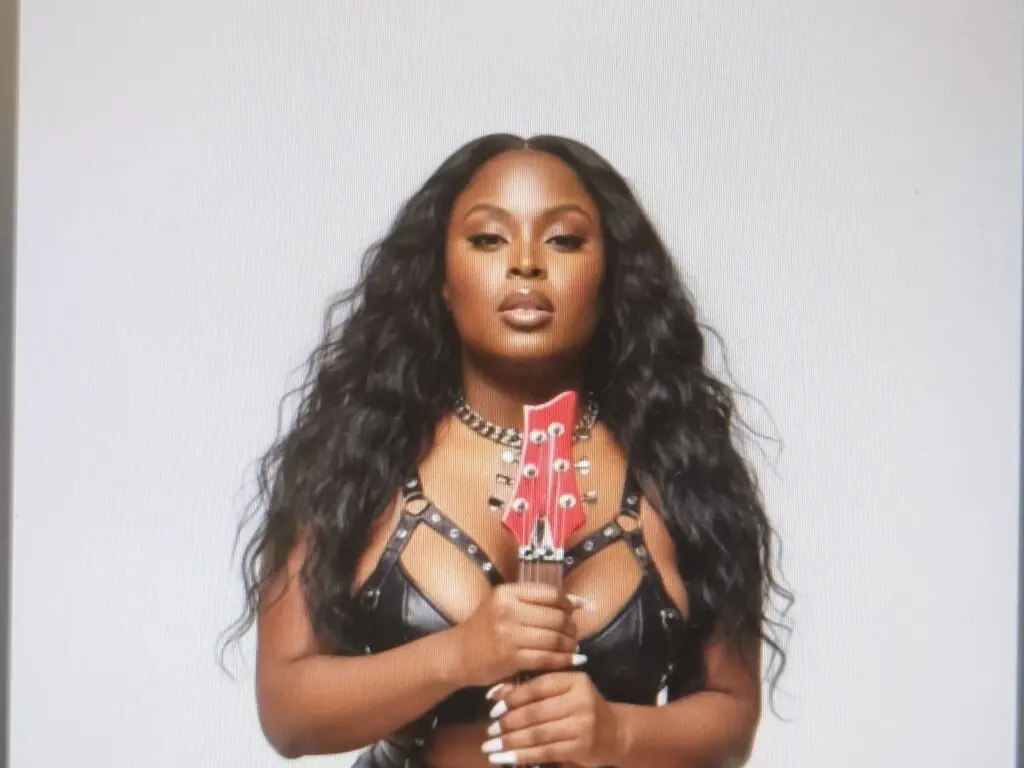 Glamorous woman with long wavy hair wearing a black leather outfit holding a red electric guitar against a white backdrop.