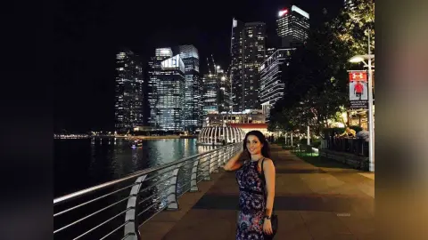 Smiling woman in a floral dress walking along a city waterfront boardwalk at night with illuminated skyscrapers.