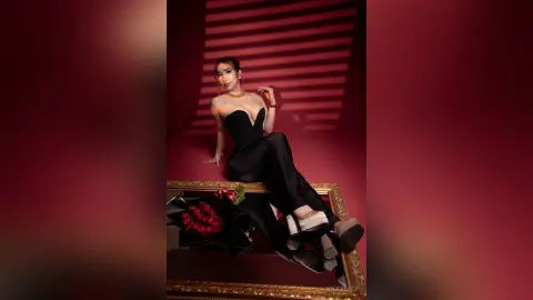Elegant woman in a black evening jumpsuit and gold jewelry posing against a moody red background.