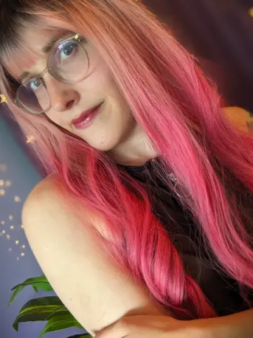 Smiling woman with long pink hair and glasses wearing a black top in a soft, dreamy indoor setting.