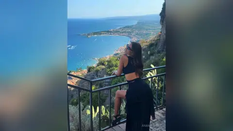 AnaMathers stands on a balcony overlooking the ocean, wearing a black crop top and skirt with her hair down.