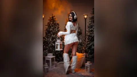 Smiling woman in white winter outfit holding a lantern amidst snowy Christmas trees in a cozy festive setting.