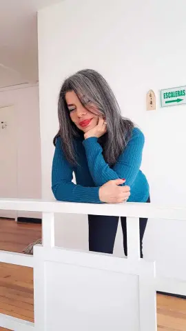 Woman in a teal ribbed sweater leaning on a white railing in a bright indoor setting.