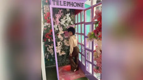 Young woman in a beige sweatshirt and brown flared pants posing in a whimsical floral telephone booth.