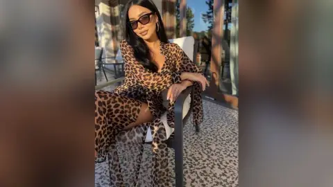 Stylish woman in a leopard print maxi dress and sunglasses sitting on a sunny outdoor patio.