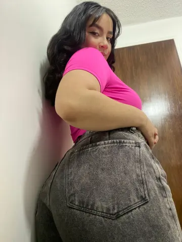 Curvy woman in a pink t-shirt and grey denim jeans posing against a white wall in a casual indoor setting.