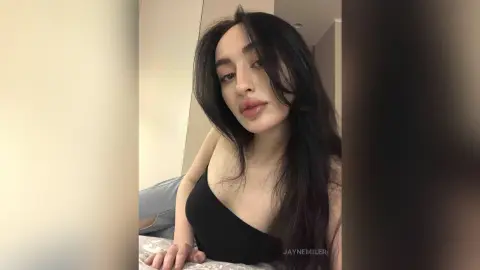 Young woman with long dark hair wearing a black tank top in a bright, minimalist indoor setting.
