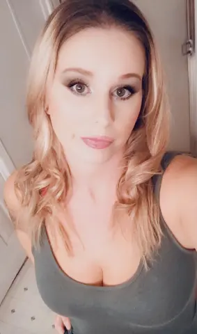 Blonde woman in a grey tank top posing for a casual indoor selfie with a soft, natural look.