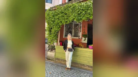 Stylish blonde woman in a black jacket and white top standing by a lush green vine wall in a sunny courtyard.