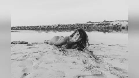 Smiling woman in a bikini relaxing on a sandy beach by the ocean in a serene black and white portrait.