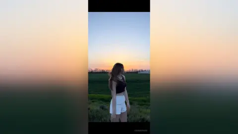 Young woman in a black crop top and white shorts standing in a green field during a golden sunset.