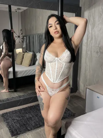 BellaFloyd poses in a white lace corset and matching thong, showing off her long dark hair and tattoos in a bedroom setting.