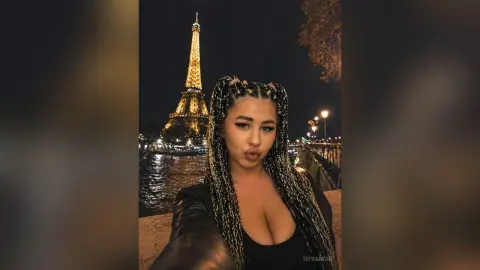 Young woman with long braids in a black outfit posing at night in front of the illuminated Eiffel Tower in Paris.