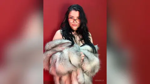 Glamorous woman with dark hair and glasses wearing a luxurious faux fur coat against a red background.