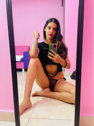 Cattalenoxx poses in a black crop top and black thong while sitting on a tiled floor against a pink wall.