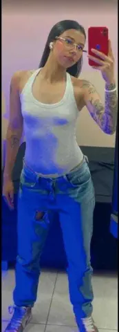 Young woman in white tank top and blue jeans taking a mirror selfie in a casual indoor setting.
