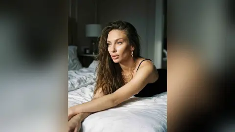 Moody portrait of a woman with long wavy hair wearing a black tank top and gold necklaces on a white bed.
