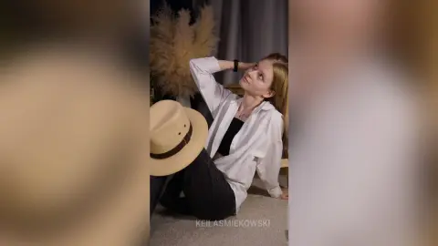 Blonde woman in white button-down shirt and black pants sitting relaxed in a cozy, minimalist room.