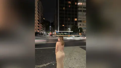 Elegant woman in a beige backless evening gown standing on a city street at night near urban buildings.