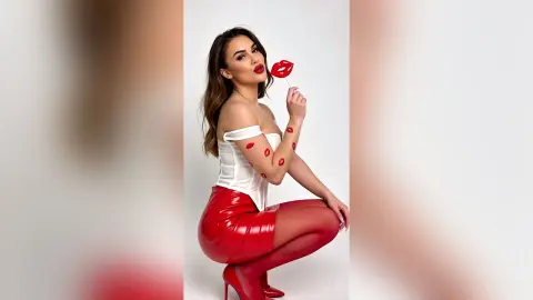 Glamorous woman in a white corset and red leather skirt holding a red lip lollipop against a white background.